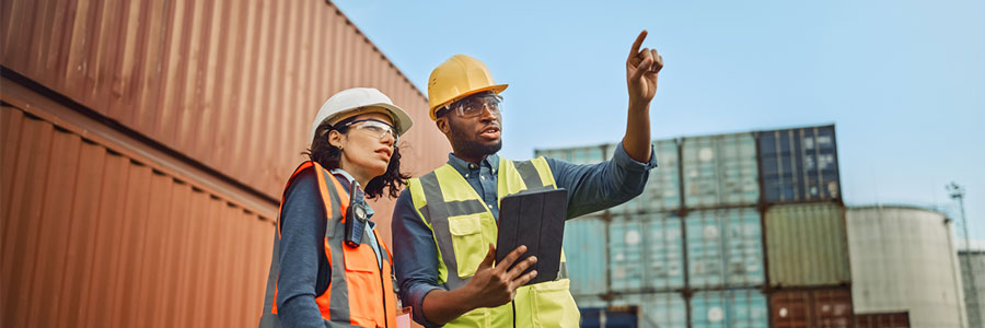 A man and a woman wearing safety helmets and high-visibility vests at a shipping container yard